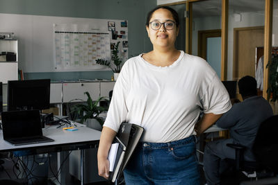 Portrait of smiling female business professional standing with hand on hip at coworking office