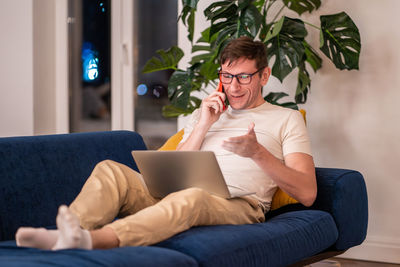 Smiling positive man freelancer talks phone works on laptop sits on couch at home in evening.