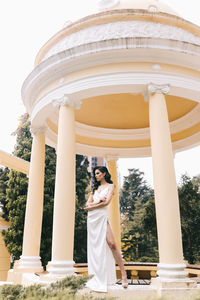 A beautiful brunette lady in an elegant wedding dress poses among the columns in the old city park