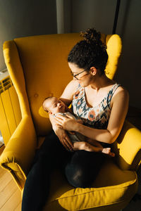 Woman sitting on yellow sofa at home holding her new born baby