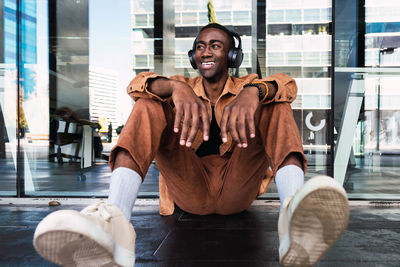 Stylish african american male listening to music in wireless headphones and looking away while sitting near glass wall in city