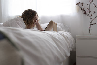 Woman using a tablet computer lying on bed in white bedroom.