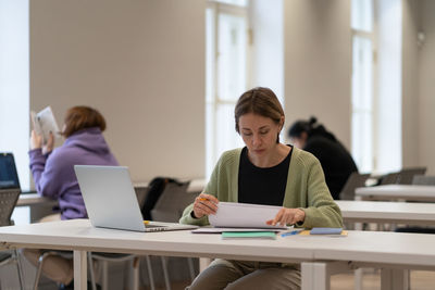 Hard-working female mature student reading architectural drawings while studying in public library