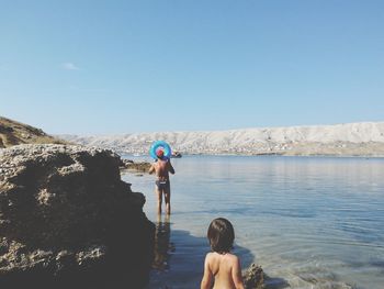 Shirtless father and son standing at riverbank against clear blue sky