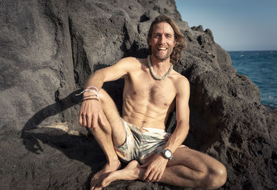 Portrait of smiling shirtless man sitting on rock at beach