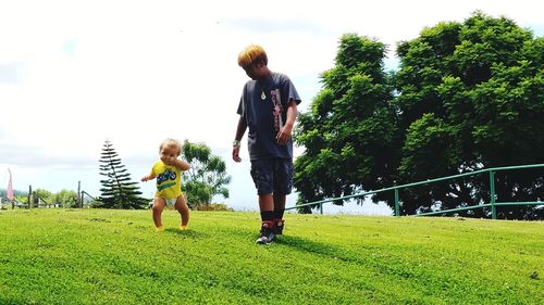 Siblings playing with ball on grass against sky