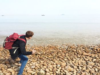 Side view of young man with backpack skimming stones in sea against sky