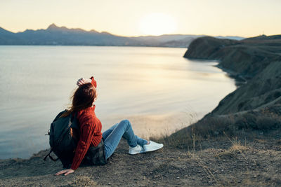 Woman sitting on rock at shore against sky during sunset