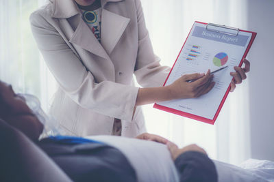 Midsection of businesswoman showing graph to colleague lying on bed in hospital