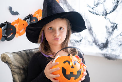 Little girl in costume of witch holding pumpkin jack with candies, celebrating halloween at home