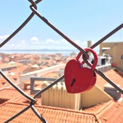 Close-up of red heart shape padlock on chainlink fence against townscape