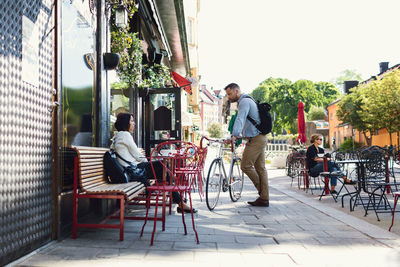 Full length of businessman with bicycle talking to colleague at sidewalk cafe