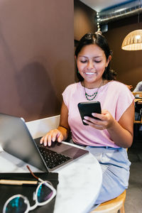 Happy young ethnic self employed lady with long dark hair in casual clothes smiling while messaging on mobile phone sitting at table with laptop during remote work in cafe