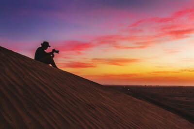 Low angle view of man photographing on sand dunes against sky during sunset