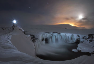 Lonely unrecognizable traveler with glowing lantern standing on rock near small frozen lake with waterfalls among desert snowy terrain at sunset time in iceland