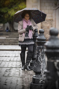 Woman holding umbrella while using phone by railing in city