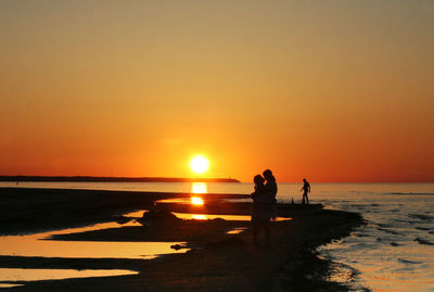 Woman with daughter standing on shore at beach against sky during sunset