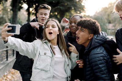 Cheerful teenage friends taking selfie through mobile phone while standing on street in city