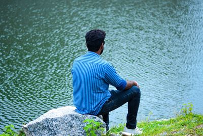 Rearview of a young man sitting on rock beside the water.