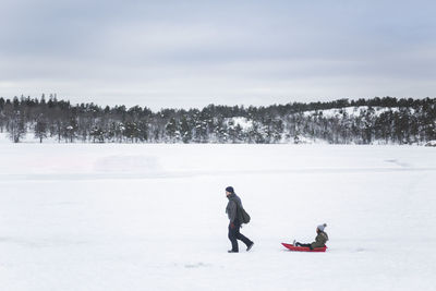 Full length of man pulling daughter sitting on toboggan while walking during winter