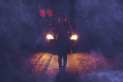 Rear view of teenage girl holding balloons while standing by illuminated car at night