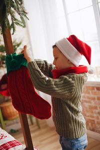 Side view of boy wearing santa hat holding stocking while standing at home