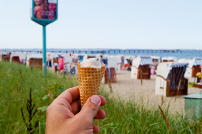 Cropped hand holding ice cream against hooded chairs at beach