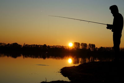 Silhouette man standing by lake against sky during sunset