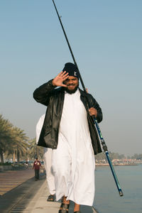 Portrait of happy man with fishing rod walking on promenade against sky