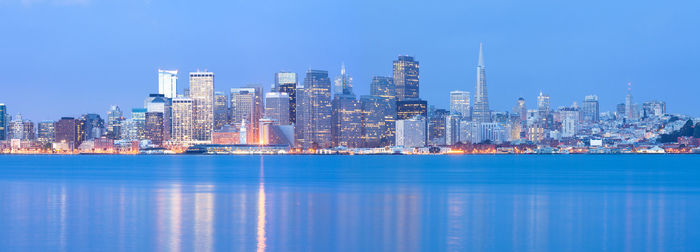 Illuminated modern buildings by bay against blue sky