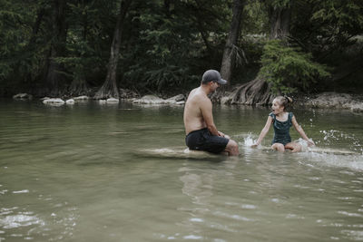 Shirtless father talking with daughter while sitting on rocks in lake against trees at forest