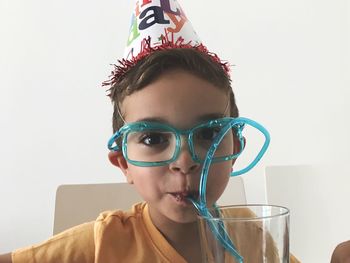 Portrait of boy in party hat having drink through strawglasses against wall at home