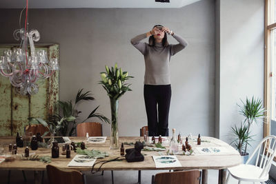 Young woman standing on chair photographing perfume bottles over table in workshop
