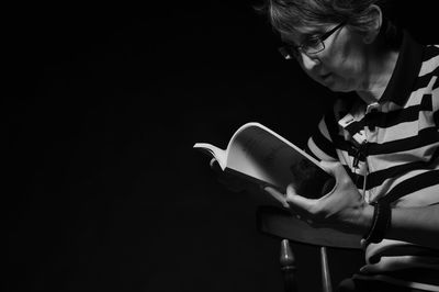 Senior woman reading book while sitting against black background