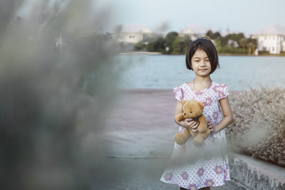 Smiling girl holding teddy bear while standing against water and sky