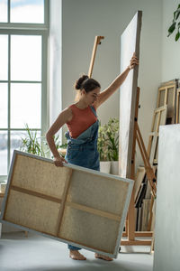 Gilr artist stands in home art studio, carrying large canvas in hands. working process in art studio