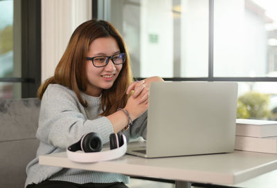Teenage girl wearing glasses with laptop and headphone on the table is chatting with smiling friend.