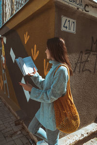 Woman reading book while standing by wall