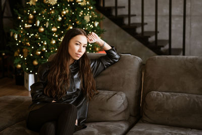 Caucasian happy woman in leather shirt with long sleeves sits on gray sofa