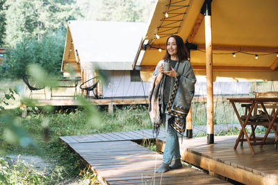 Brunette woman in poncho drinking tea and relaxing in glamping in nature