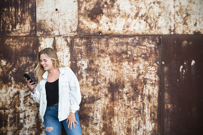 Young woman using phone while standing against wall