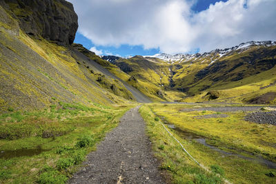 Pathway amidst volcanic landscape leading towards mountain range against sky