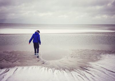 Rear view of girl walking at beach against cloudy sky during winter