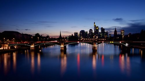 Illuminated bridge over river by buildings against sky at night