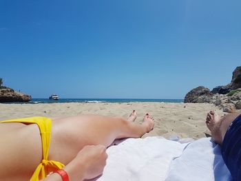 Low section of couple relaxing at beach against clear blue sky on sunny day