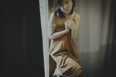 Young woman looking away while standing against window at home