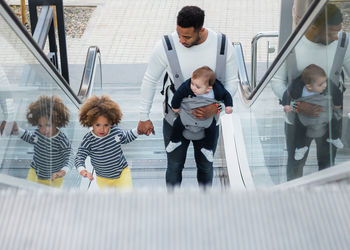 From above of positive young ethnic man carrying infant son and holding hand of cheerful little daughter while standing on escalator in city