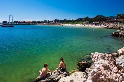 People sitting on rocks by sea against sky