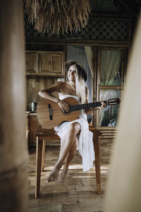 Full length of smiling woman playing guitar sitting at home