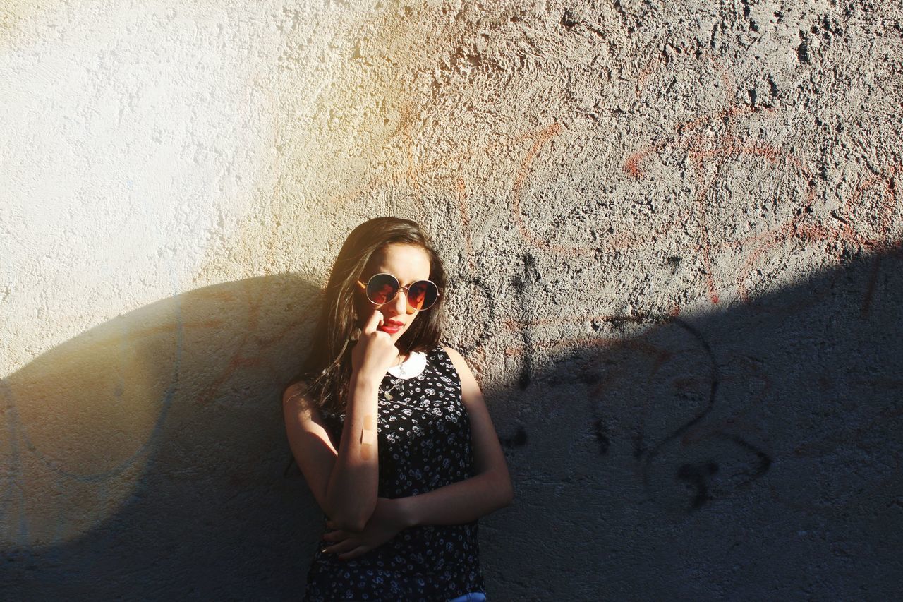 PORTRAIT OF YOUNG WOMAN WEARING SUNGLASSES STANDING IN SUNLIGHT